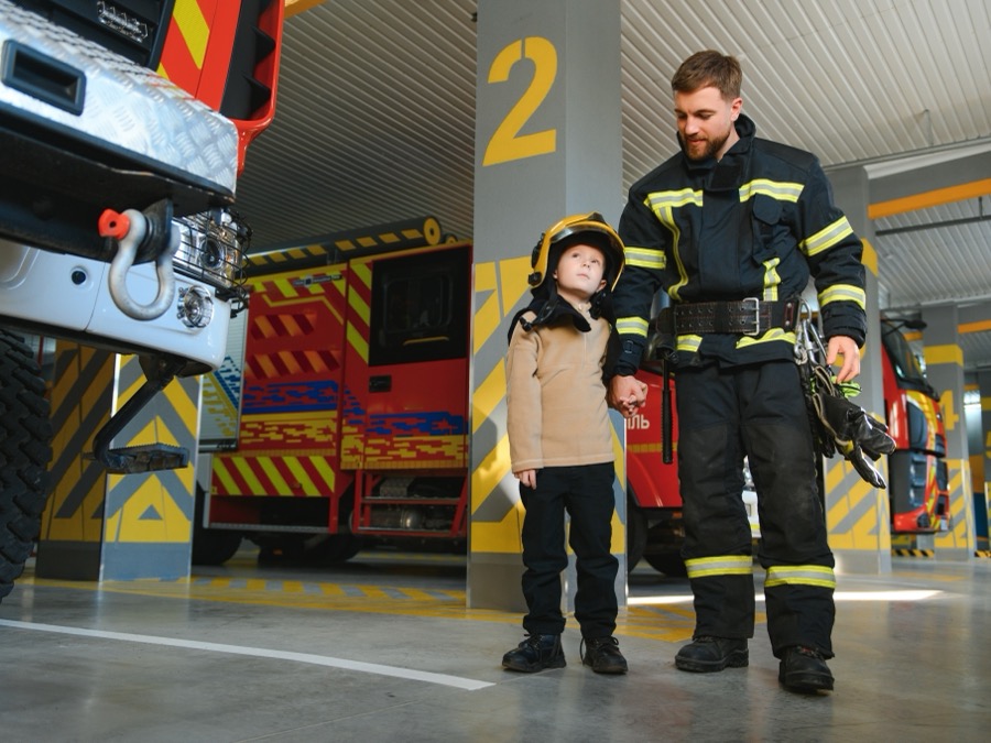 Firefighter holds a young boy's hand walking through a fire station garage bay lined with red engines