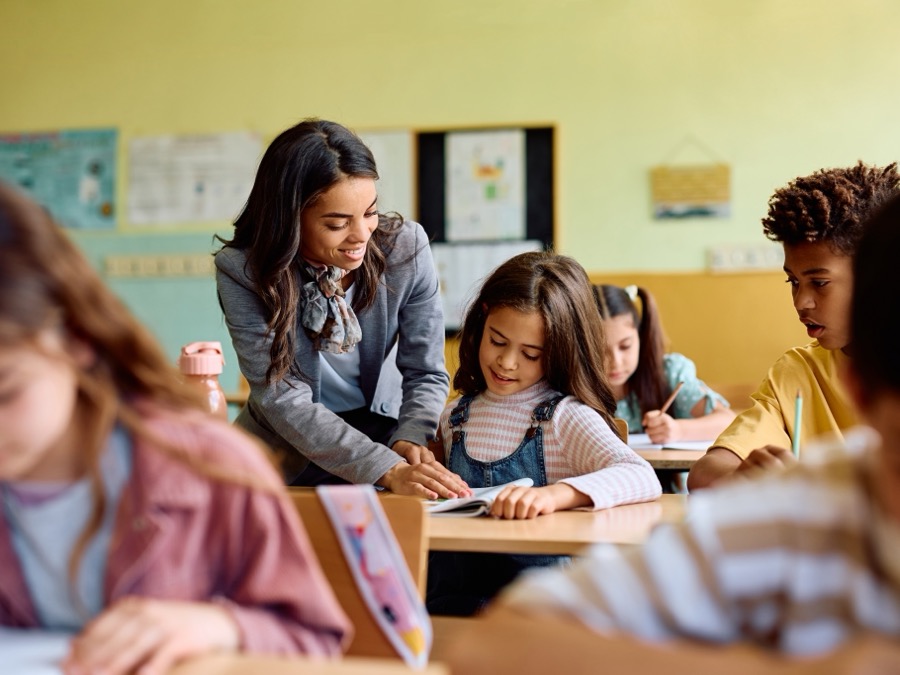 Smiling teacher leans over a desk to help a young girl with schoolwork in a sunlit elementary classroom