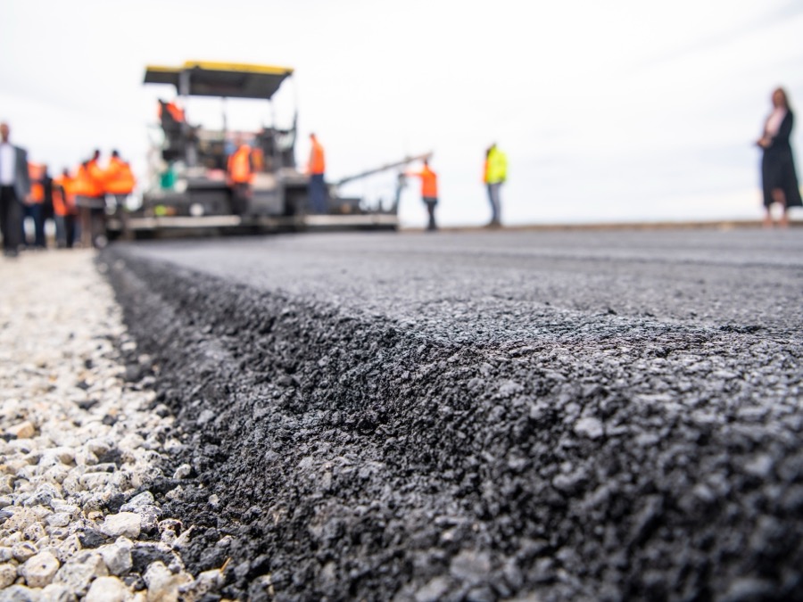 Close-up of a freshly paved asphalt road edge meeting a gravel shoulder, paving crew blurred behind