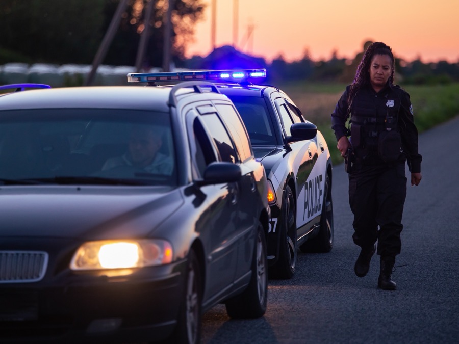Female police officer walks along a rural roadside at dusk near her marked patrol car and a stopped vehicle