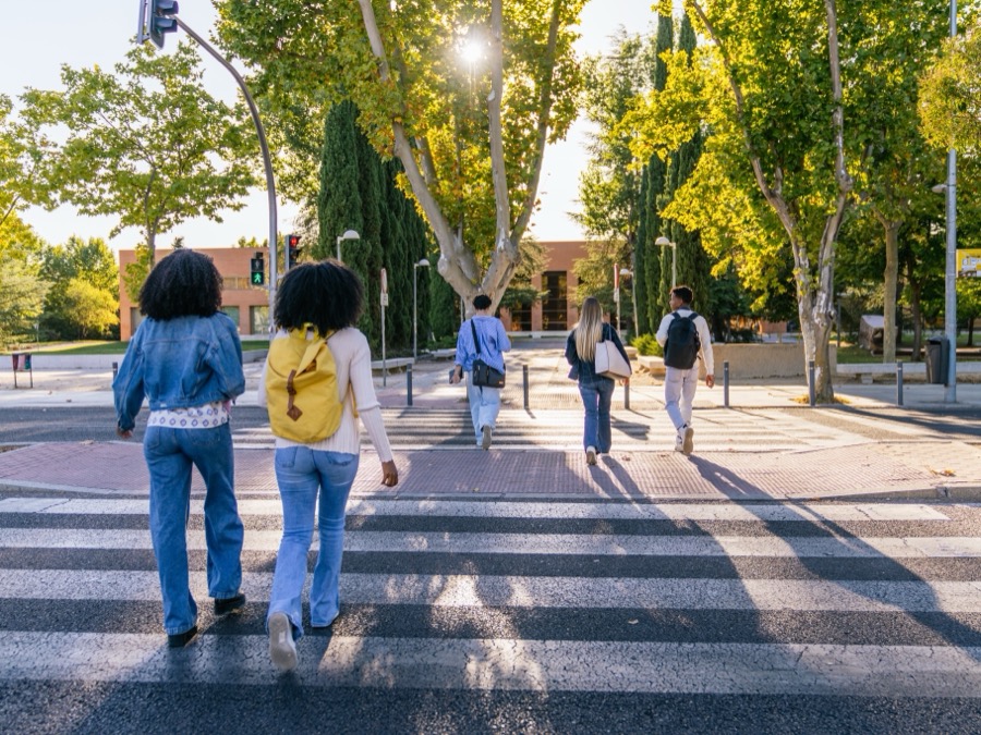 Five students with backpacks cross a striped crosswalk toward a sunlit, tree-lined campus plaza