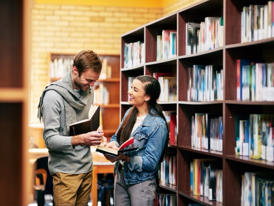 A man and woman smile in a library aisle, each holding an open book, beside tall wooden shelves