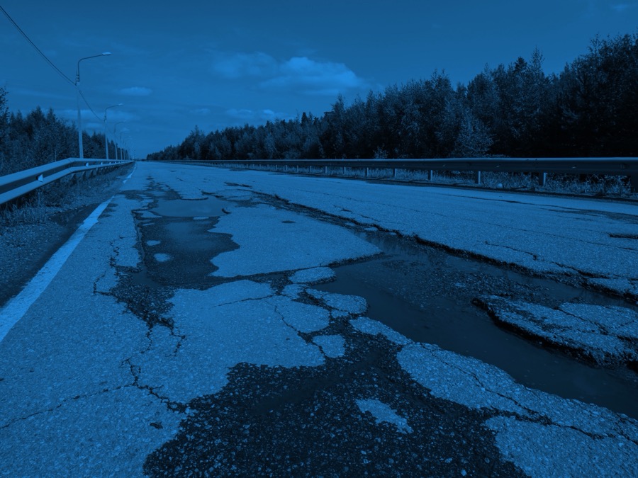 Deteriorated rural highway with wide cracks, crumbling edges, and pooled water under an overcast sky